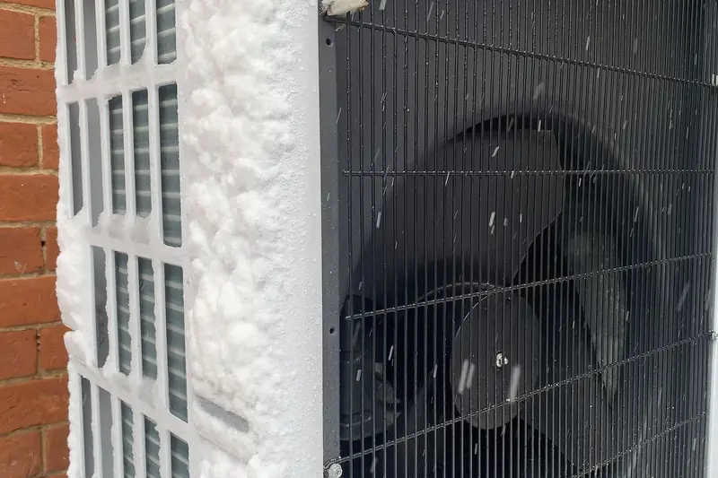 Close view of a heat pump's outdoor fan grille beside a red-brick wall, its side panel caked thick with snow