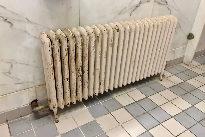 A long cream-colored cast-iron radiator with paint flaking off its columns, standing on a gray and white checkered tile floor