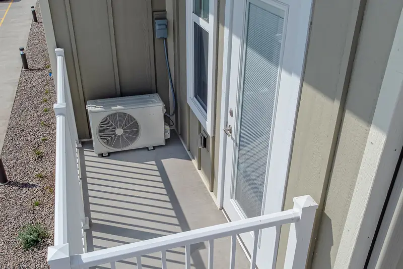 Compact white outdoor condenser sitting on a gray upstairs balcony beside a glazed door, white railings in front
