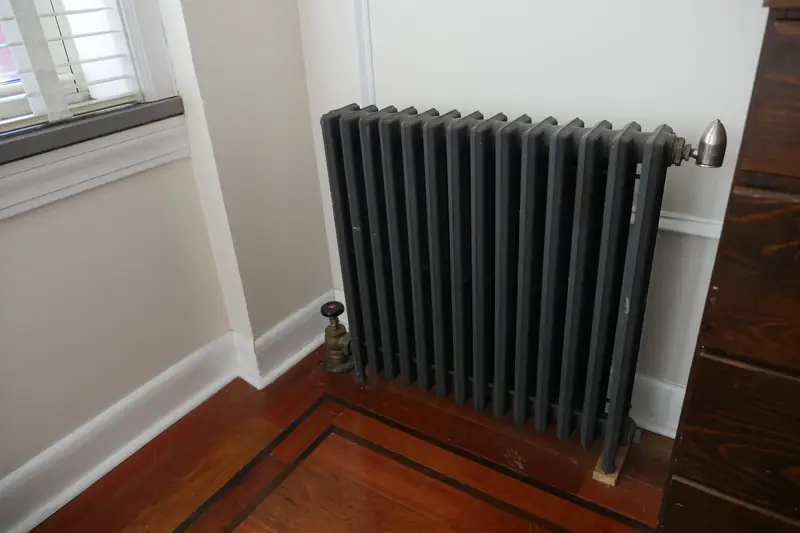 Charcoal-painted cast-iron steam radiator in a white corner by a window, a brass supply valve at the floor on one end and a small bullet-shaped vent at the top of the other